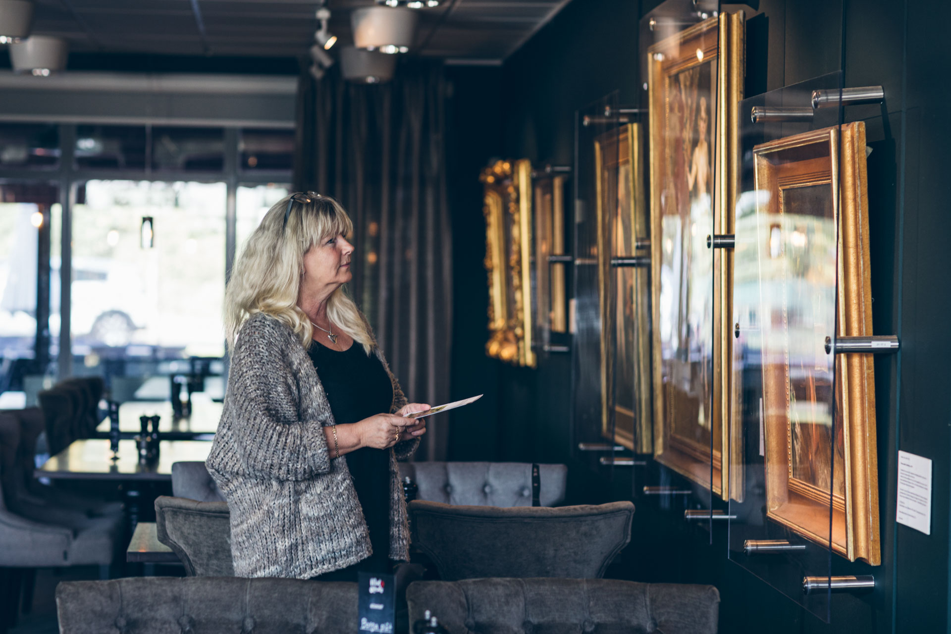 Kvinna med blont hår och grå kofta betraktar tavlor i konstgalleri eller museum